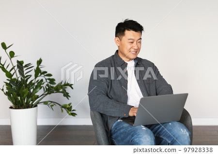Smiling middle aged chinese male sitting in armchair, texting, typing on computer, surfing in internet Smiling middle aged chinese male sitting in armchair, texting, typing on computer, surfing in internet 97928338