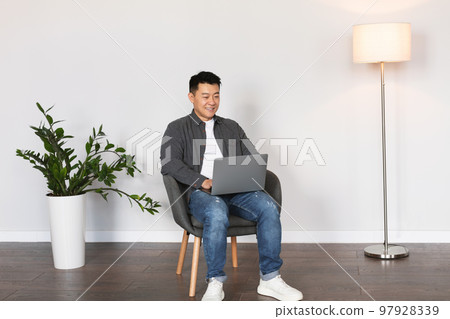 Cheerful middle aged korean man typing on laptop surfing in internet, chatting in living room Cheerful middle aged korean man typing on laptop surfing in internet, chatting in living room 97928339
