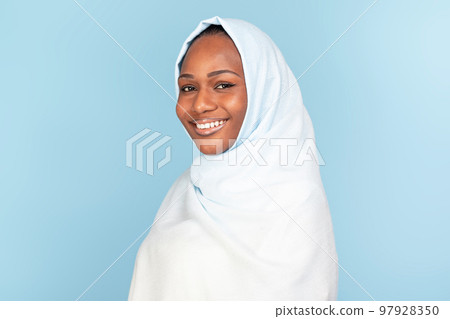 Portrait of beautiful black muslim woman in hijab posing on blue studio background, smiling at camera, copy space 97928350
