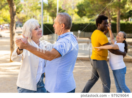 Exited older and middle-aged diverse people enjoying latin dances breaking into pairs on sandy area between trees 97929064