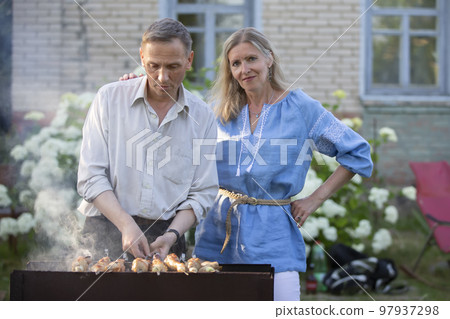 An elderly couple, a man and a woman, are preparing a barbecue. 97937298