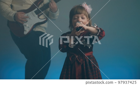 Little girl on stage in vintage dress, she sings into microphone and dances, her father plays an electric guitar. Color music is shining and smoke is billowing. Family time, creativity and hobbies 97937425