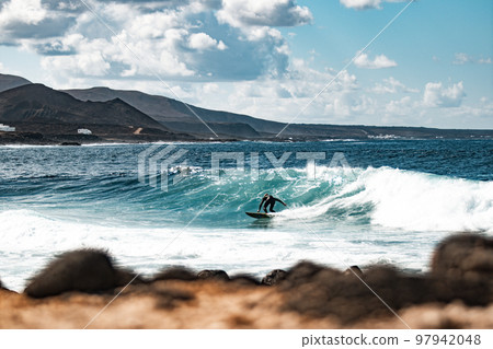 Wild rocky coastline of surf spot La Santa Lanzarote, Canary Islands, Spain. Surfer riding a big wave in rocky bay, volcano mountain in background 97942048