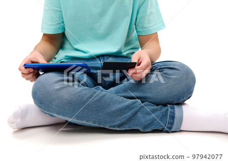 Cropped view of a teenage boy, wearing blue jeans and turquoise t-shirt, sitting in lotus pose on white background, using mobile app on his smartphone and credit card, purchasing online. Copy ad space Cropped view of a teenage boy, wearing blue jeans and turquoise t-shirt, sitting in lotus pose on white background, using mobile app on his smartphone and credit card, purchasing online. Copy ad space 97942077