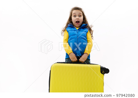 Surprised baby girl with long curly hair in blue jacket, carrying a heavy yellow suitcase, expressing surprise, astonishment, posing with open mouth, looking at camera, isolated on white background. 97942080