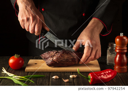 Chef hands slicing beef steak with knife on wood cutting board. Food preparation process concept. Haute cuisine Chef hands slicing beef steak with knife on wood cutting board. Food preparation process concept. Haute cuisine 97942254
