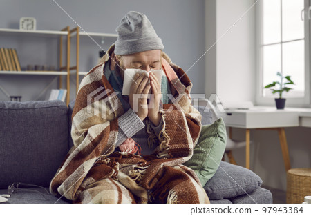 Old man is sitting at home, warmly wrapped in warm blanket, dressed in knitted sweater and hat. 97943384