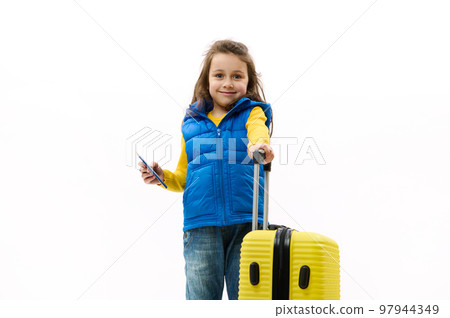 Caucasian lovely little kid girl, wearing a blue jacket, jeans and yellow pullover, smiles looking at camera, posing with a travel bag and boarding pass on white background. Tourism Travel. Copy space 97944349