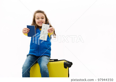 Mischievous overjoyed child girl sitting on yellow suitcase, holding out at camera passport and air ticket, rejoicing at upcoming travel abroad, isolated over white background. Copy advertising space 97944350