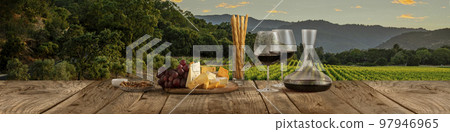 Decanter and two glasses with delicious red wine, grape and cheese appetizers standing on wooden table over beautiful nature landscape of fields and forest. Decanter and two glasses with delicious red wine, grape and cheese appetizers standing on wooden table over beautiful nature landscape of fields and forest. 97946965