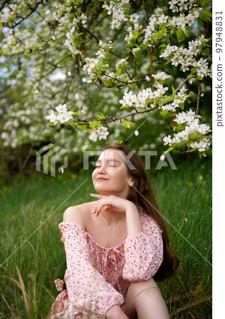 The girl is standing under a tree with her eyes closed. White petals are falling on the face The girl is standing under a tree with her eyes closed. White petals are falling on the face 97948831
