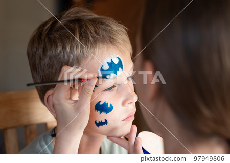 Child animator, artist's hand draws face painting to little boy. Child with funny face painting. Painter makes blue bat mask on boy's face. Child animator, artist's hand draws face painting to little boy. Child with funny face painting. Painter makes blue bat mask on boy's face. 97949806