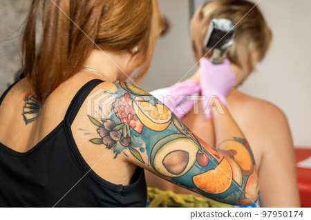 Young woman with sleeve tattoo at the work. Tattoo session at studio. Back view. Defocused client on the background. Close up 97950174