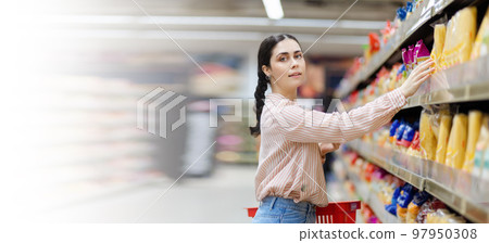 Web banner of shopping. Portrait of young smiling beautiful caucasian woman reaches hand to takes food from shelf. Mock up. Copy space. Aisle in background. Concept of consumerism Web banner of shopping. Portrait of young smiling beautiful caucasian woman reaches hand to takes food from shelf. Mock up. Copy space. Aisle in background. Concept of consumerism 97950308