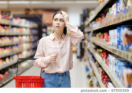 Portrait of young shocked caucasian woman holds cart and takes hand to head. Showcase at background. Person forget to buy something. Concept of shopping in supermarket and consumerism Portrait of young shocked caucasian woman holds cart and takes hand to head. Showcase at background. Person forget to buy something. Concept of shopping in supermarket and consumerism 97950309