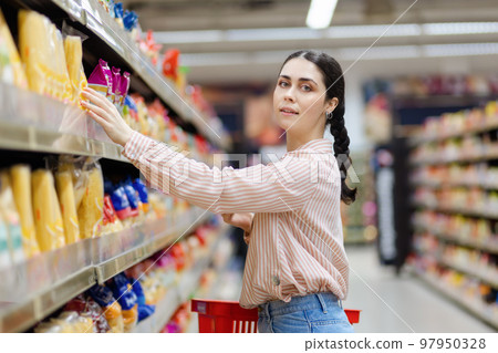 Portrait of young smiling beautiful caucasian woman reaches to top shelf. Aisle in background. Concept of shopping and consumerism 97950328