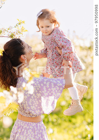 a mother and little daughter in a lilac dresses on a walk in the spring garden.  a mother and little daughter in a lilac dresses on a walk in the spring garden.  97950998
