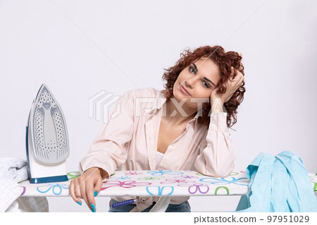 The girl leans her elbow against the ironing board and her hand against her head. 97951029