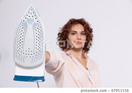 A woman with curly hair holds an iron in her hand and looks into the camera. 97951556