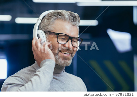 Close-up photo portrait of mature gray-haired businessman, gray-haired man in headphones listening to music audio books and podcasts online, boss at workplace inside office. Close-up photo portrait of mature gray-haired businessman, gray-haired man in headphones listening to music audio books and podcasts online, boss at workplace inside office. 97951821
