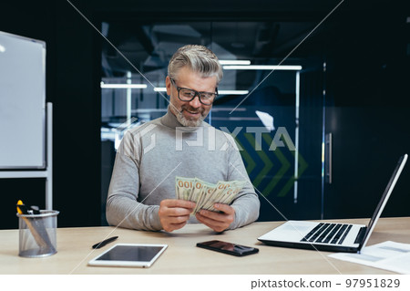 Mature successful financier investor counts money dollars cash sitting inside office at table, senior businessman happy with result achievement and income uses laptop at work. Mature successful financier investor counts money dollars cash sitting inside office at table, senior businessman happy with result achievement and income uses laptop at work. 97951829
