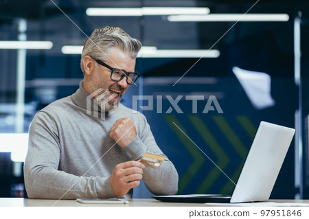 Happy and smiling businessman in office making bank transfer and online purchase in online store, mature man holding bank credit card and using laptop, happy with choice. Happy and smiling businessman in office making bank transfer and online purchase in online store, mature man holding bank credit card and using laptop, happy with choice. 97951846