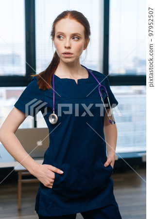 Vertical portrait of thoughtful female practitioner in medical uniform looking away standing on background of window. Woman physician with hands at waist posing with stethoscope at hospital. Vertical portrait of thoughtful female practitioner in medical uniform looking away standing on background of window. Woman physician with hands at waist posing with stethoscope at hospital. 97952507