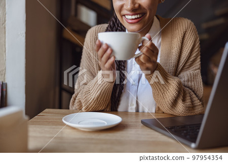 Focus on beautiful toothy smile of pretty ethnic woman holding cup with cappuccino during work on laptop in coffee shop Focus on beautiful toothy smile of pretty ethnic woman holding cup with cappuccino during work on laptop in coffee shop 97954354