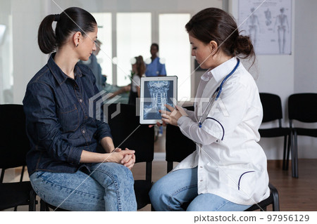 Female medical specialist interpreting digital x ray in hospital waiting room. Doctor explaining young patient diagnosis in medical clinic. Woman listening explanation of sanatorium nurse. 97956129