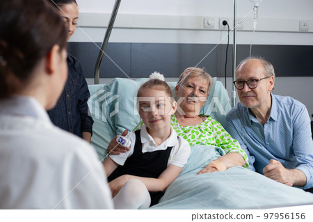 Elderly patient, family members listening attentively instructions of unrecognizable female health professional. Surgeon physician visiting grandmother bedridden in sanatorium room. Elderly patient, family members listening attentively instructions of unrecognizable female health professional. Surgeon physician visiting grandmother bedridden in sanatorium room. 97956156