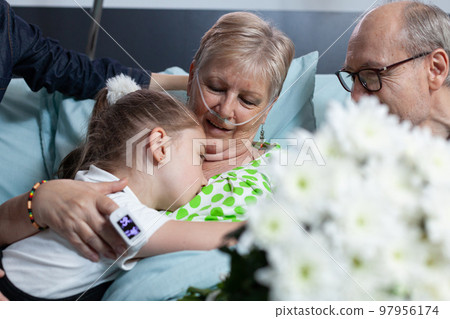 Little girl waving goodbye to sick grandmother. Elderly woman lying in hospital bed connected to medical monitoring devices hugging granddaughter. Relatives visiting old ill lady in geriatric clinic. 97956174