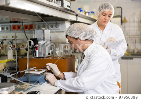 Latin male scientist using microscope while female chemist taking notes in lab 97956389