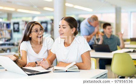 Two friendly adult female students studying in university library, using books and laptop while preparing for lecture 97956536