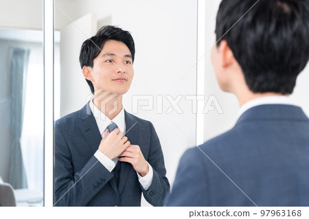 一個年輕的商人看著洗手間的鏡子，係緊領帶（早上，準備工作） 97963168