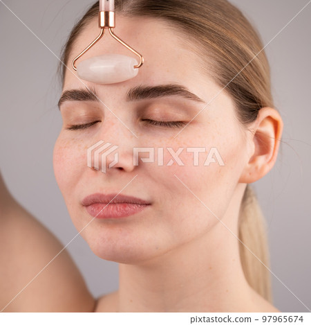 Close-up portrait of a woman uses a quartz roller massager to smooth wrinkles on her forehead. 97965674