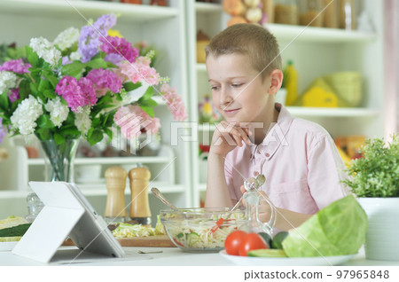 little boy eating salad and looking at tablet screen on kitchen table at home 97965848