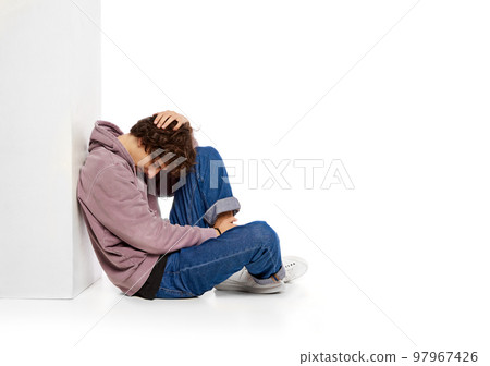 Portrait of young boy sitting on floor in depression and sadness over white background. Problems of youth Portrait of young boy sitting on floor in depression and sadness over white background. Problems of youth 97967426