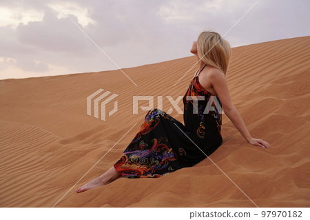 Beautiful blonde woman in a black dress is sitting on the hot sand in the desert Beautiful blonde woman in a black dress is sitting on the hot sand in the desert 97970182