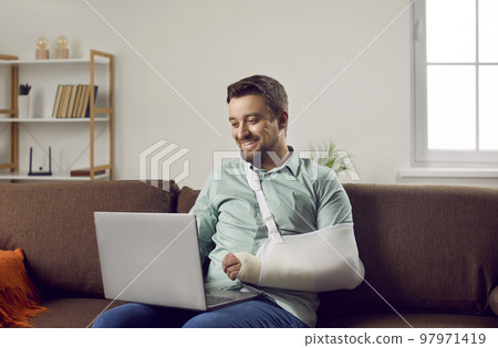 Happy young man with arm in a cast sitting on the couch at home and communicating on a laptop. Happy young man with arm in a cast sitting on the couch at home and communicating on a laptop. 97971419