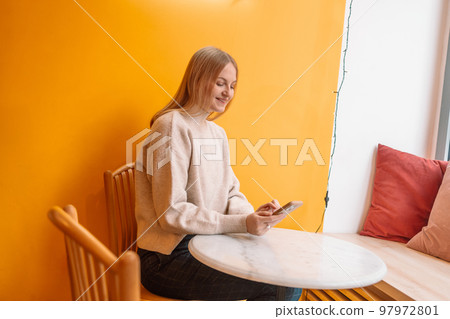 Attractive young blonde smiling woman with cup of coffee using mobile phone in cafe on a yellow background Attractive young blonde smiling woman with cup of coffee using mobile phone in cafe on a yellow background 97972801