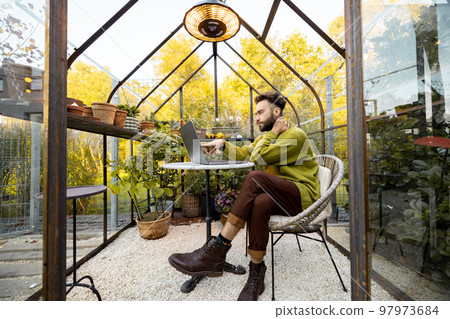 Man works remotely on laptop in glasshouse at backyard Man works remotely on laptop in glasshouse at backyard 97973684