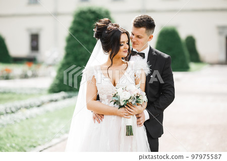 Beautiful bride and groom embracing and kissing on their wedding day outdoors Beautiful bride and groom embracing and kissing on their wedding day outdoors 97975587