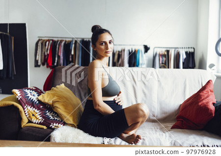 Cheerful young brunette latin pregnant sportswoman sitting on couch in light room 97976928