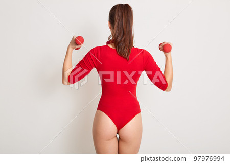 Back view of slender well built sportswoman wearing bright red combidress, holding two dumbbells in both hands, having ponytail, standing isolated over white background in studio. Sport concept. Back view of slender well built sportswoman wearing bright red combidress, holding two dumbbells in both hands, having ponytail, standing isolated over white background in studio. Sport concept. 97976994