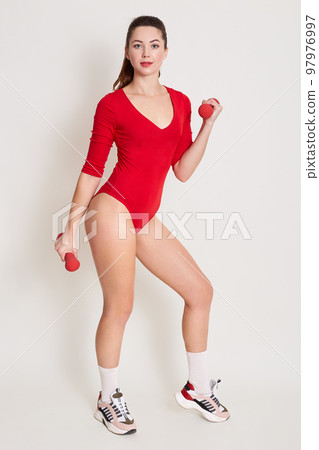 Full length portrait of gorgeous adult sports woman doing exercises for biceps with dambbells, sporty female wearing red stylish combidress, looking directly at cameras, looks confident and strong. 97976997