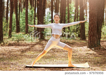 Winsome female in white sportswear practicing yoga in green park or forest, standing in yoga position, keeping eyes closed, spreading hands aside, training outdoor. 97976998