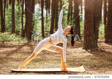 Portrait of woman in white stylish sport top and leggins standing on mat in yoga position in beautiful forest, stretching body, practicing yoga outdoors. 97976999