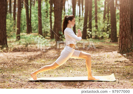 Profile full length photo of young adult woman with ponytail dresses white stylish sportswear doing yoga outdoors, pressing palms together, meditation and relaxing. 97977000