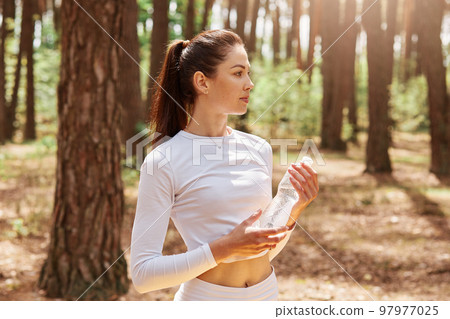 Fitness beautiful woman with dark hair and ponytail holding bottle of water and looking away, posing after exercising in forest. Female athlete in white top work out outdoor. 97977025