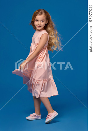 Fashionable little girl in a pink dress is posing on a blue background. 97977033